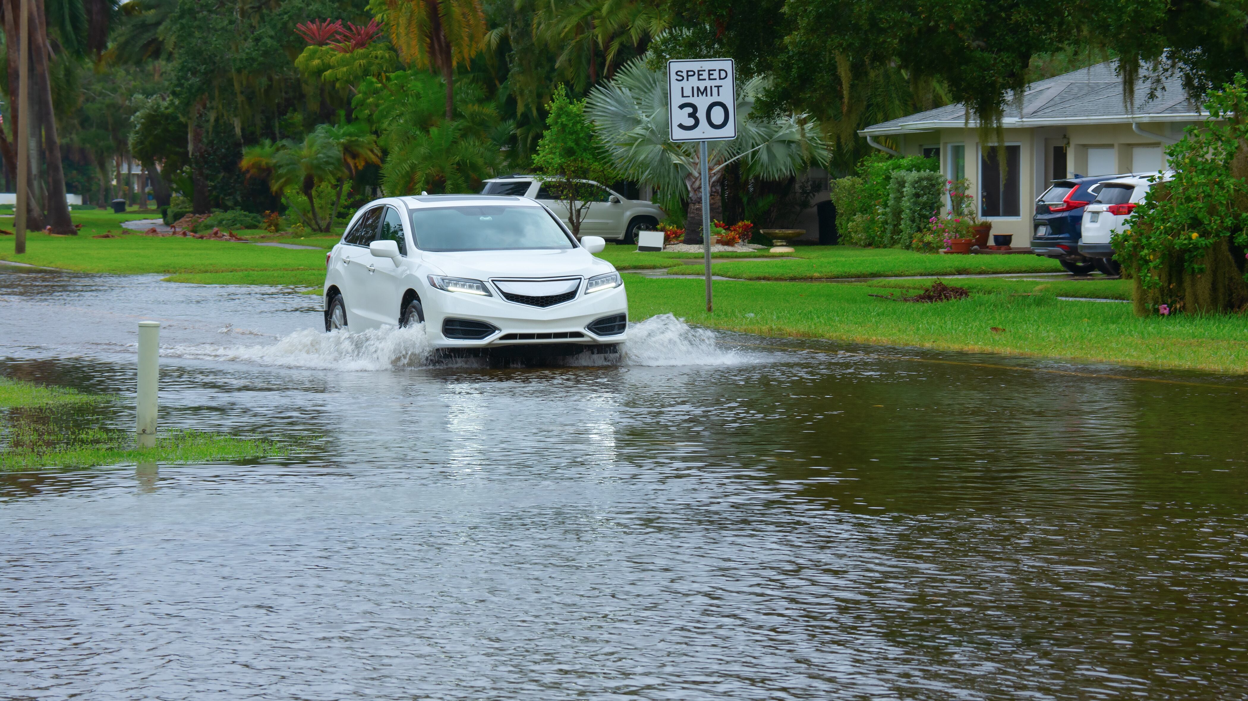 Heavy flooding and storm surge in residential neighborhood with a car driving through deep splashing water in the flooded street in front of houses with Speed Limit sign on side of the road.